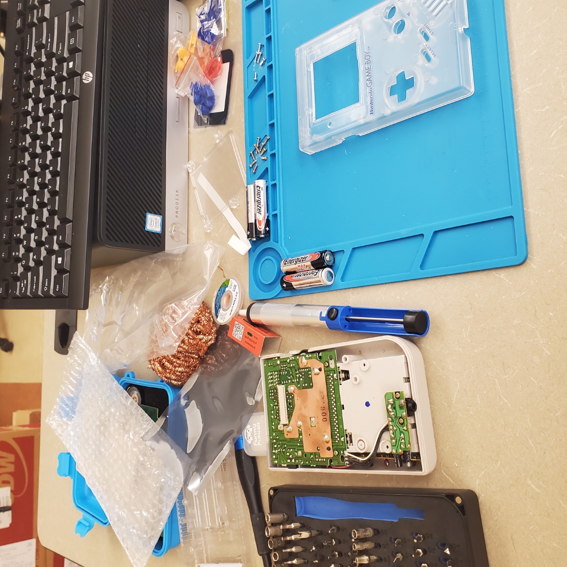 beige table with a blue workmat on it. on the workmat is a clear white gameboy shell, batteries, screws, screwdrivers, and soldering tools